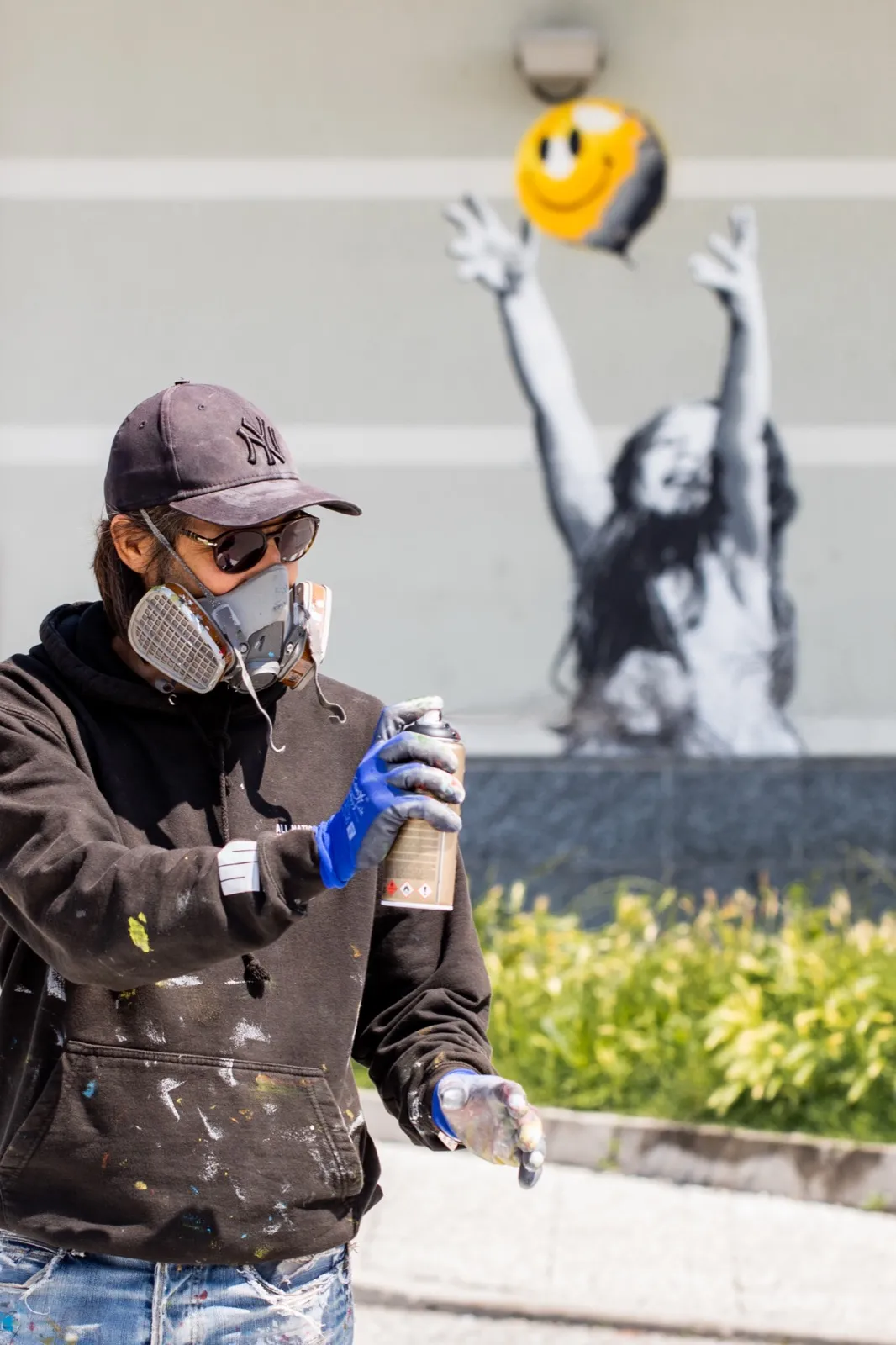 Alessio-B con maschera, occhiali e cappellino, bomboletta in mano, davanti a un murale